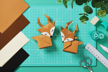 Two origami deer made of brown paper on a turquoise cutting mat. Nearby are items for paper crafts.
