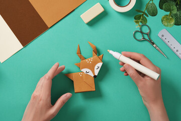 Hands draw white dots with a marker on a paper reindeer craft. Around them are paper craft supplies.