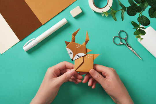 Hands holding a finished paper deer craft. Scissors, tape, glue, and colored paper.