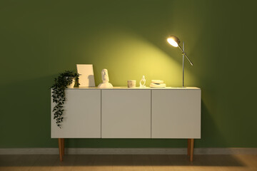 Glowing lamp on chest of drawers near green wall