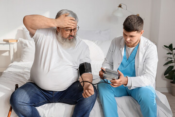 Young caregiver measuring blood pressure of ill senior man in bedroom