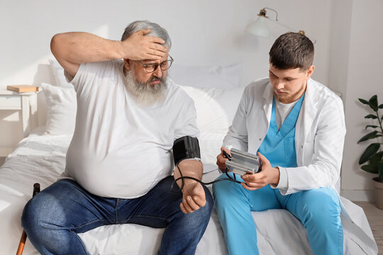 Young caregiver measuring blood pressure of ill senior man in bedroom