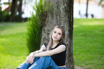 A young, beautiful blonde woman, wearing jeans and a black shirt, sits on the grass, leaning her...