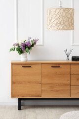 Vase with lilacs on wooden chest of drawers and lamp in stylish room