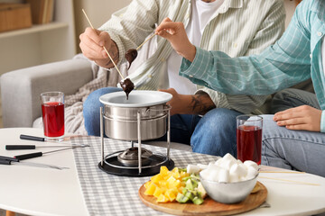 Young couple with glasses of wine dipping marshmallows into chocolate fondue at home, closeup