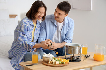 Young happy couple with glasses of juice dipping fresh oranges into chocolate fondue at home
