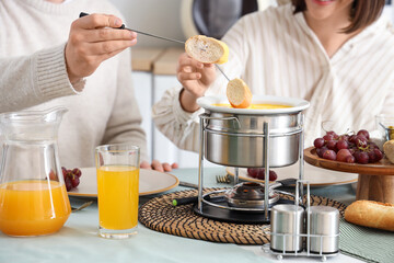 Young couple with glasses of juice dipping croutons into cheese fondue at home, closeup