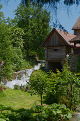 A picturesque wooden house stands beside a cascading waterfall, surrounded by lush green trees and vibrant foliage under a clear blue sky, evoking a sense of tranquil nature.