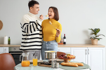 Young happy couple with glasses of juice eating croutons dipped in cheese fondue at home