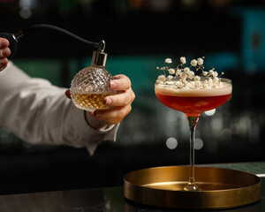 A bartender's hand is delicately misting a blush-toned, floral-garnished cocktail with a vintage...