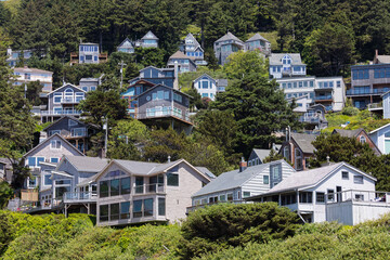 Beachfront photo of hillside homes along the Oregon Coast, seen from Tunnel Beach in Oceanside, with the Pacific Ocean, green forest, and clear blue sky on a sunny summer day