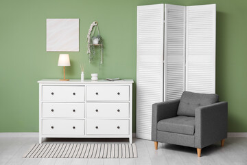 Interior of modern room with folding screen, chest of drawers and armchair near green wall