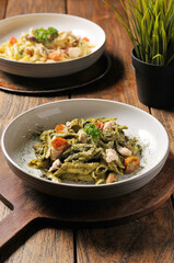 Two bowls of pasta are presented on a rustic wooden table, with one featuring green pesto pasta with chicken and cherry tomatoes in the foreground, and another bowl of pasta blurred in the background