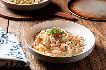 A bowl of creamy pasta with chicken and diced red bell peppers is centrally placed on a wooden table, accompanied by a patterned napkin and a blurred pan in the background.