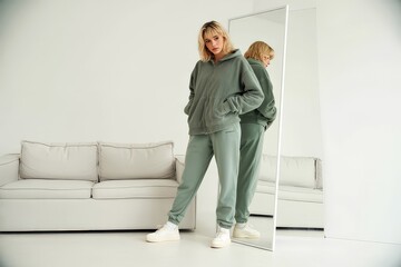 A young woman models a comfortable green hoodie and sweatpants set in a minimalist home setting