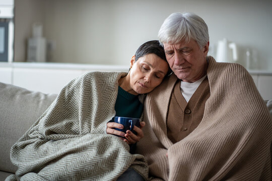 Sad elderly couple hug under blanket with hot tea, both suffering from illness at home. Infection quarantine, shared sickness, family weakened condition, domestic isolation, senior health struggle