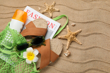String bag with bottle of sunscreen cream, magazine and beach accessories on sand as background,...