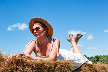 Young beautiful sexy woman in a straw hat and sunglasses lying on a haystack on a meadow on a sunny day against the sky. Rest. Vacation. Tourism. Romance. Countryside.