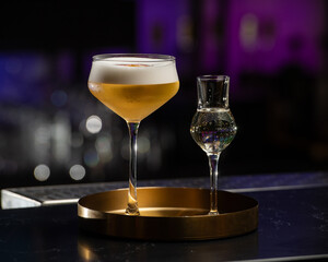 A beautifully presented golden-hued cocktail with a rich foam top is served alongside a small shot glass of clear liquid, all resting on a chic golden tray
