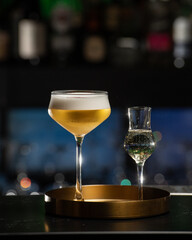 A beautifully presented golden-hued cocktail with a rich foam top is served alongside a small shot glass of clear liquid, all resting on a chic golden tray