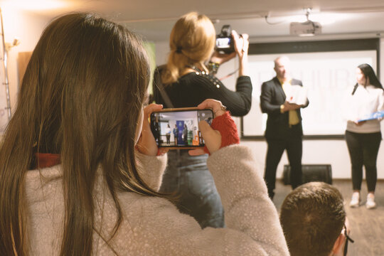 Audience member films a presentation on stage using a smartphone, capturing an event with speakers and a screen.