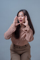 Young Indonesian woman in beige shirt, holding her hands near her face with a playful expression,...