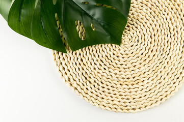 Top view of brown wicker table mat, green monstera leaf on white background. Straw round tray, flat lay, copy space. 