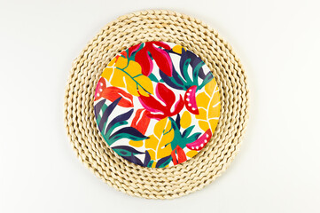 Top view of brown wicker table mat, colorful plate on white background. Straw round tray, exotic plant plate, summer picnic. Flat lay, copy space. 
