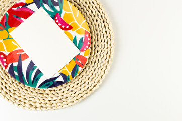 Top view of brown wicker table mat, white card, colorful plate on white background. Straw round...