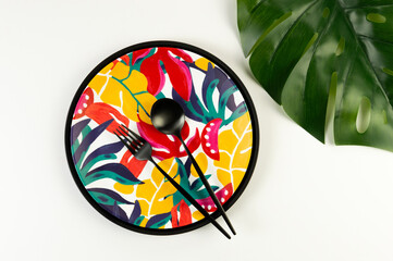 Top view of black tray, colorful plate, black cutlery on white background. Exotic plant plate, monstera palm leaf, summer picnic. Flat lay, copy space.