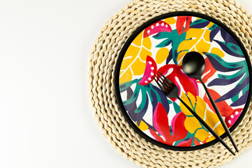 Top view of brown wicker table mat, colorful plate, black cutlery on white background. Black tray, straw round tray, exotic plant plate, summer picnic. Flat lay, copy space. 