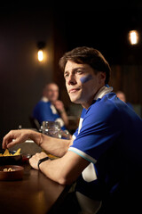 Caucasian young man sitting at table watching soccer match, with face painted in team colors,...
