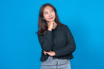 Portrait of a young Asian woman thinking while standing against a blue background. She looks up...