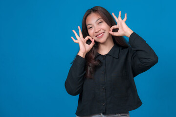 Happy young Asian woman smiling and showing double OK hand gestures near her face while wearing a...