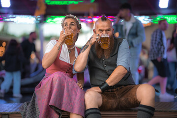 Smiling couple in Bavarian traditional costume holding beer mugs at a lively folk festival in...