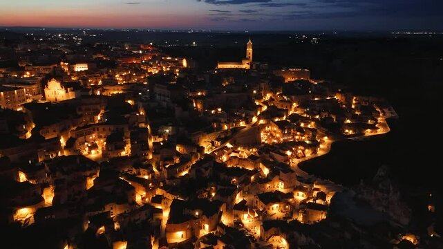Sassi of Matera, historic UNESCO old town glowing under amber street lights. Night time drone aerial