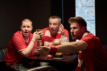 Three anxious Caucasian soccer fans sitting at table watching match, holding drinks, reacting with...