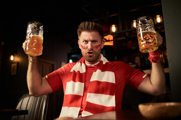 Caucasian young man with face paint and striped scarf cheering while holding two large beer mugs in...