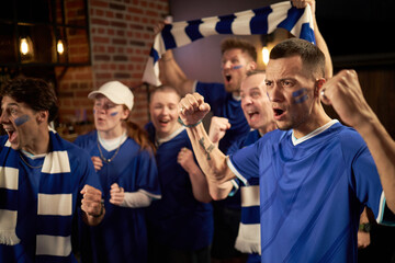 Fototapeta premium Group of diverse young soccer fans cheering and celebrating soccer match victory in championship, wearing blue jerseys and scarves, showing excitement and team spirit