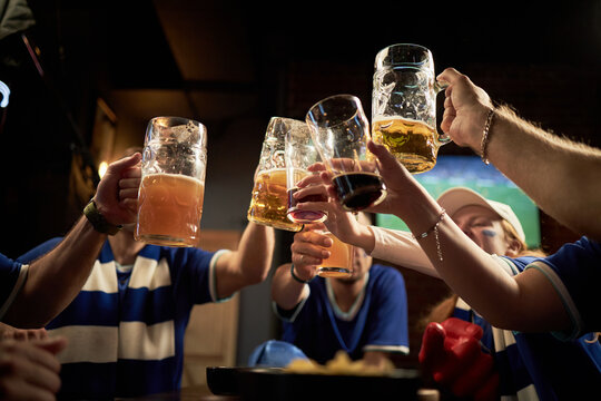 Group of young soccer fans raising beer mugs and toasting in pub in celebration, wearing team jerseys, sitting together in sports bar, watching live match on television