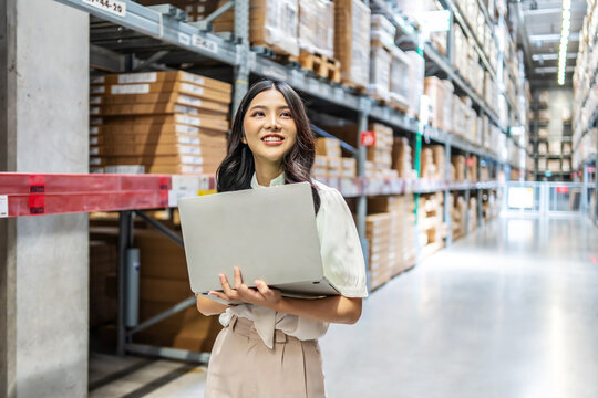 Engaging in meticulous inventory management, a young asian woman professional examines products while using a laptop computer, surrounded by organized shelves filled with boxes