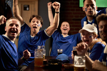 Group of multiethnic young men and women celebrating goal while watching soccer match in sports bar, raising arms and cheering with excited expressions, drinks and snacks on table