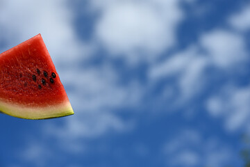 red slice of watermelon held against a blue sky with scattered clouds