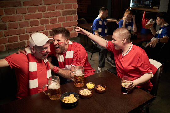 Group of young Caucasian fans in red jerseys celebrating and cheering while watching soccer match in sports bar, holding beer mugs and snacks on table, wearing team scarves, showing excitement