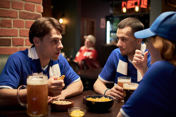 Three young Caucasian soccer fans in blue jerseys sitting at table in sports bar eating snacks and...