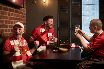 Three Caucasian sport fans including one woman and two young men celebrating soccer match in bar, wearing fan gear, holding beer mugs, smiling and cheering together at table