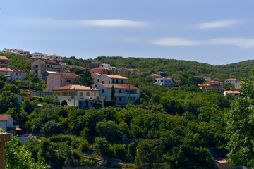 Obraz premium A vibrant cluster of houses with terracotta roofs nestled amidst a dense, green hillside, under a bright blue sky, showcasing a typical Mediterranean residential landscape.
