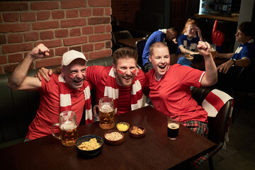 Three Caucasian young men and woman celebrating and cheering with drinks and snacks at table in sports bar, wearing soccer fan attire, smiling and raising arms, while watching football
