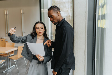 Two colleagues from diverse backgrounds thoughtfully discussing a document, collaborating in an office space with a modern interior, highlighting teamwork and professional interaction.