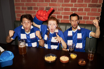 Three young Caucasian soccer fans with painted faces cheering and celebrating, while watching match together, at table with drinks and snacks in pub or sports bar